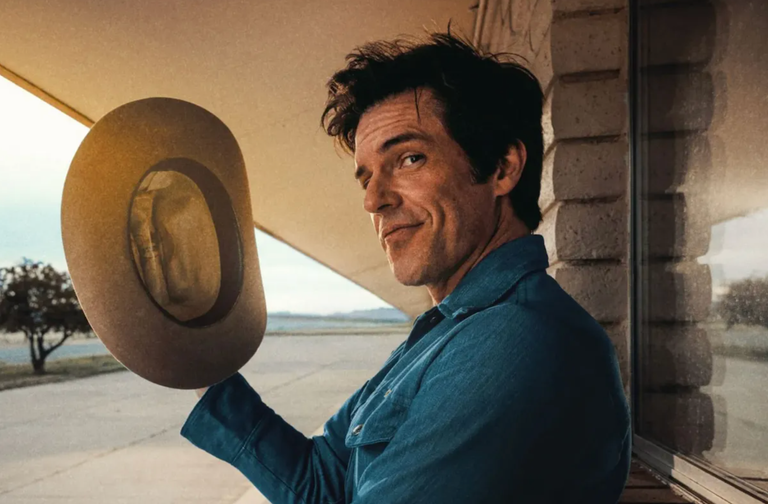 Musician Brandon Flowers smirks at the camera while tipping a light-colored cowboy hat outdoors against a brick building backdrop.